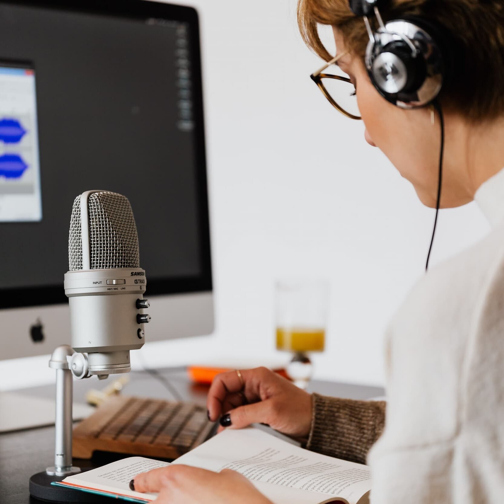 Woman wearing headset with microphone and computer monitor on her desk. She's reading from a book.