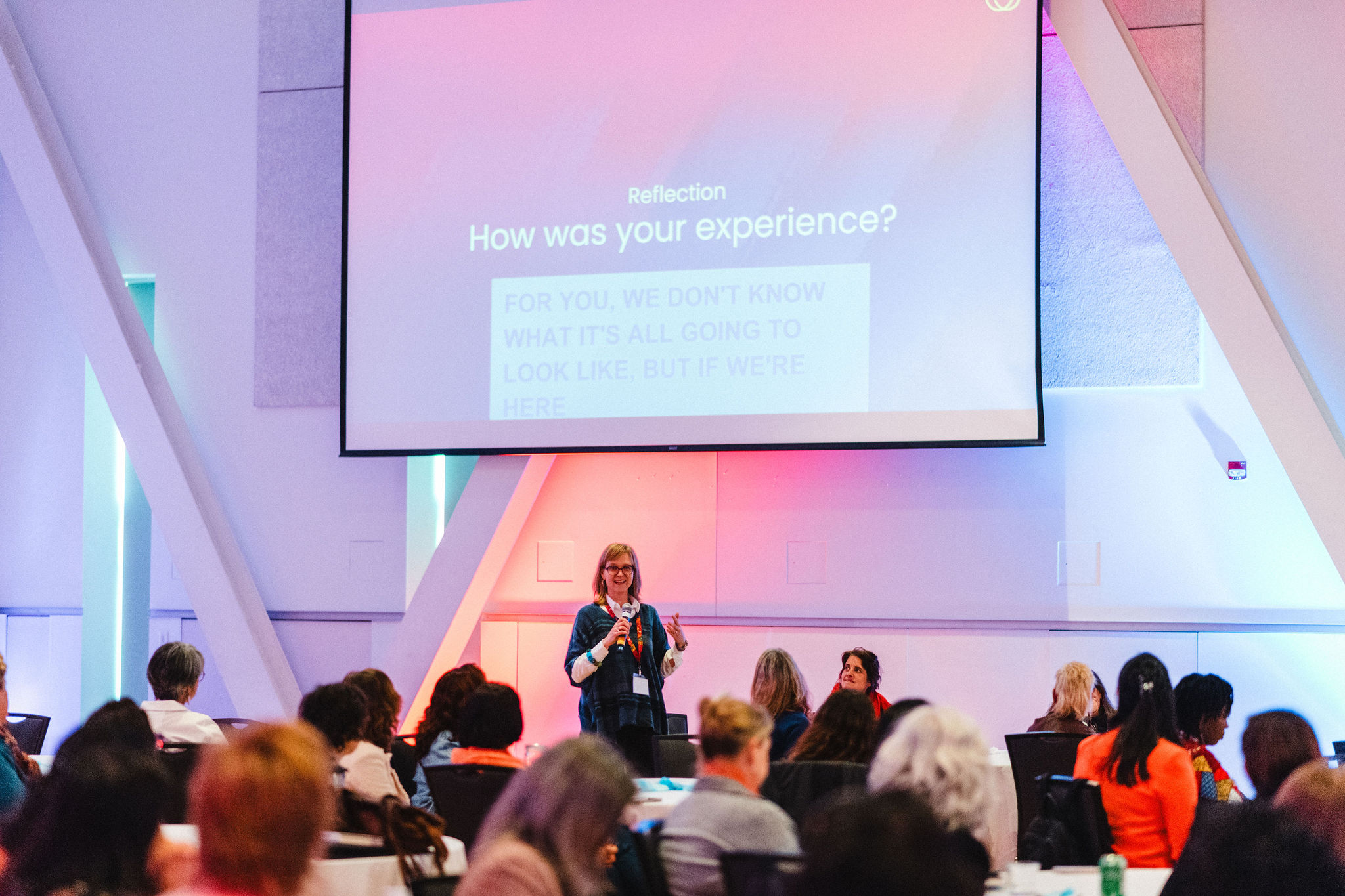 Woman on a stage speaking to a large audience. There is a large screen behind her showing captions of what she's saying.