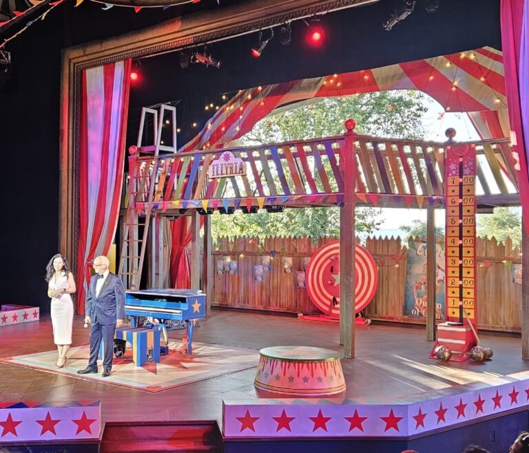 Theatre set depicting a stage decorated with a circus theme. A man and woman are standing in formal attire at the centre, with an audience in front of them.