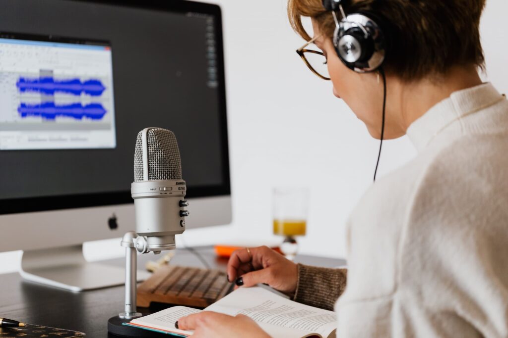 Woman wearing headset with microphone and computer monitor on her desk. She's reading from a book.